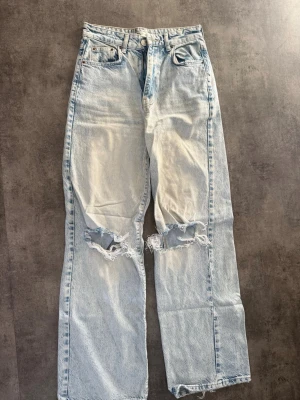Ljusblå slitna jeans från Gina Tricot - Ljusblå jeans med raka ben och hög midja från Gina Tricot. Jeansen har slitna detaljer och hål vid knäna för en trendig look. Klassisk femficksmodell i denim med dragkedja och knapp framtill.