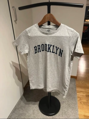 Grå t-shirt med BROOKLYN-tryck - Snygg grå t-shirt från 157 med stort BROOKLYN-tryck i mörkblått på bröstet. Klassisk rund halsringning och korta ärmar. Perfekt för en avslappnad streetstyle-look.