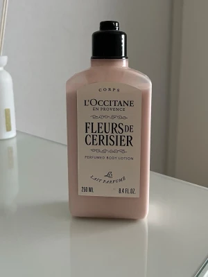 L'Occitane Fleurs de Cerisier Body Lotion - L'Occitane Fleurs de Cerisier är en parfymerad body lotion i en ljusrosa plastflaska med svart pump. Innehåller 250 ml och har en söt doft av körsbärsblom. 