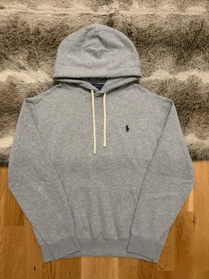 Grå Polo Ralph Lauren Fleece Hoodie - Storlek M - Hej! 👋 Säljer en klassisk ljusgrå Polo Ralph Lauren hoodie i storlek M med marinblå pony-logga. 🐎 Tröjan är i mycket gott skick, helt utan hål, fläckar eller andra defekter. Riktigt välvårdad och fräsch! 100% äkta med originaltvättlappar kvar och en fullt fungerande QR-kod för superenkel verifiering ✅ Det absolut bästa och mest tidlösa basplagget man kan ha i garderoben. Fraktar snabbt! 🚛📦💬💯 