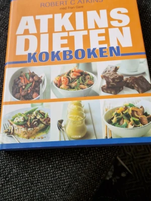 Atkinsdieten Kokboken - En färgstark kokbok med hårda pärmar, skriven av Robert C. Atkins. Omslaget är orange och blått med flera matbilder som visar olika rätter. Perfekt för dig som vill laga mat enligt Atkinsdieten och få inspiration till nya lågkolhydratrecept.