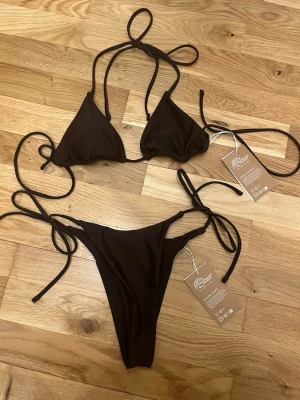 Brun bikini med knyt från nakd - Snygg brun bikini från Palm Collective med klassisk trekantsmodell på överdelen och knyt i sidorna på både topp och trosa. Enkel att justera för bästa passform. Helt ny aldrig använd. 