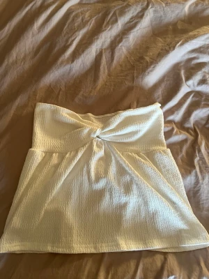 Vit bandeau tubtopp med twist - Snygg vit bandeau tubtopp med twistdetalj framtill. Toppen är ärmlös och har en lätt strukturerad yta i ett mjukt material. Perfekt för en trendig och fräsch look i sommar.