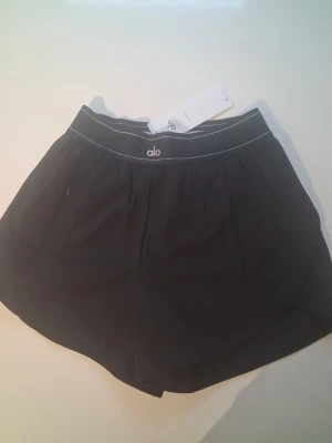 Svarta träningsshorts från Alo - Svarta träningsshorts från Alo med elastisk midja och vit kontrastsöm. Lätta och bekväma, perfekta för aktiva dagar. Klassisk design med diskret logga framtill och praktiska fickor.