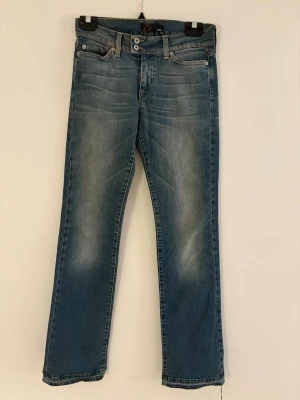 Blå raka jeans från Nudie Jeans - Säljer ett par klassiska blå jeans från Nudie Jeans med raka ben och snygg slitning framtill. Jeansen har fem fickor, dubbla knappar i midjan och kontrastsömmar på bakfickorna. Perfekta till en avslappnad stil.