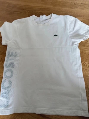  t-shirt Lacoste  - Vit t-shirt från Lacoste med klassisk krokodillogga på bröstet och stor textlogga längs sidan i mesh-tyg. T-shirten har rund halsringning och korta ärmar. Perfekt för en clean och sportig stil.