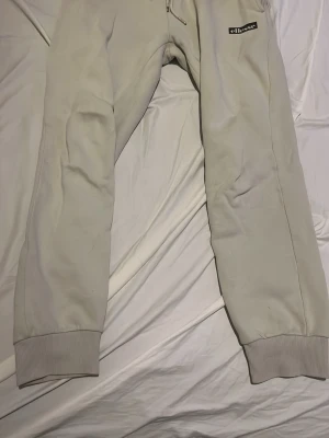 Beige mjukisbyxor från Ellesse - Snygga beige mjukisbyxor från Ellesse med resår i midjan och vid bensluten. Byxorna har en liten Ellesse-logga framtill och en bakficka med diskret etikett. Perfekta för en chill och avslappnad stil.