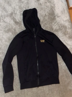 Svart hoodie från EA7 Emporio Armani - Snygg svart hoodie med dragkedja från EA7 Emporio Armani. Hoodien har huva, ribbade muddar och fickor framtill. EA7-logga i guld på bröstet ger en clean och exklusiv vibe. Tillverkad i mjukt material som känns skönt mot huden.