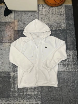 Vit hoodie med dragkedja från Lacoste - Vit hoodie från Lacoste med dragkedja och huva. Klassisk krokodillogga på bröstet och två fickor framtill. Regular fit-modell i mjukt material, perfekt för en avslappnad stil.