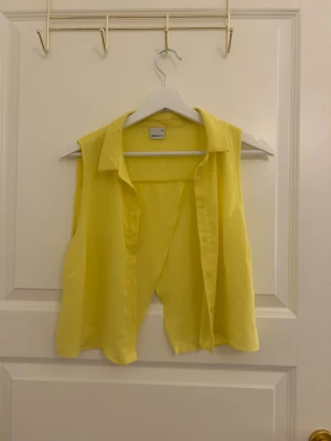 Gul ärmlös blus från Gina Tricot  - Snygg gul blus från Gina tricot  med klassisk krage och knäppning framtill. Blusen är ärmlös och har en luftig, lätt och flowy passform som passar perfekt till sommaren. Tillverkad i ett tunt och mjukt material som känns skönt mot huden.