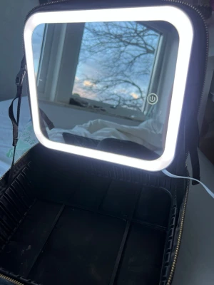 Sminkspegel med LED-belysning - Snygg sminkspegel/sminkväska med fyrkantig form och inbyggd LED-belysning som kan växla mellan kallt och varmt ljus. Spegeln sitter i ett svart fodral med dragkedja och har touch-knapp på spegeln för att styra ljuset. Perfekt för sminkning och styling hemma eller på resan. Kan dimma