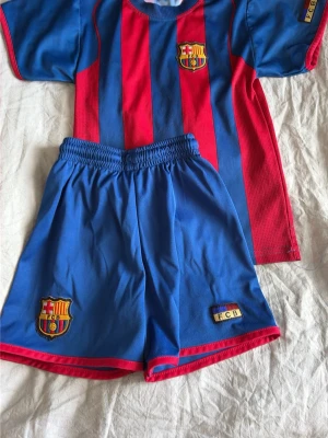 Barcelona matchställ röd/blå - Säljer ett officiellt FC Barcelona matchställ med tröja och shorts. Tröjan har klassiska röda och blå vertikala ränder och klubbmärke på bröstet. Shortsen är blå med klubbmärke och elastisk midja. Perfekt för fotbollsträning eller att bära som supporter. Storlek 128 