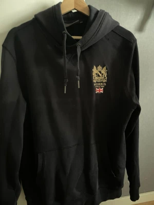 Svart hoodie från Morris med tryck - Svart hoodie från Morris med guldfärgat broderat emblem och brittisk flagga på bröstet. Klassisk huva med dragsko och känguruficka framtill. Perfekt för en stilren och avslappnad look.