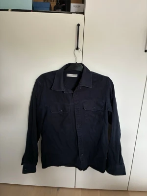 Mörkblå overshirt från James harvest - Snygg mörkblå skjorta från James Harvest med två bröstfickor och knappar framtill. Skjortan har klassisk krage och långa ärmar, perfekt för en stilren look. Tillverkad i mjuk bomull som känns skön mot huden. Nypris 800 kr!