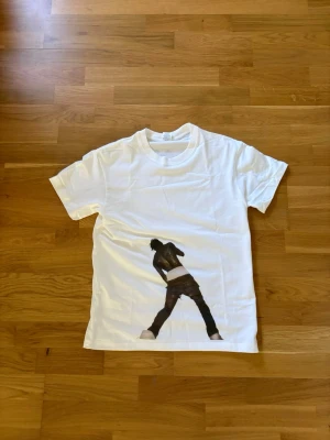 Vit t-shirt med svart tryck - Vit t-shirt med en stor svart siluett-tryck av en person på framsidan. Klassisk rund hals och korta ärmar. Perfekt för en avslappnad och cool stil.