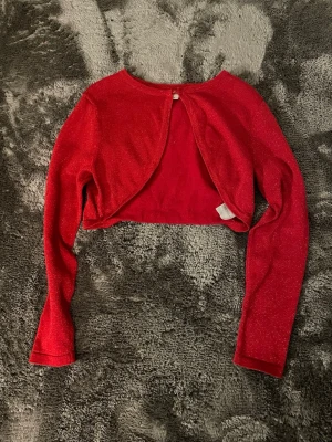 Röd glittrig bolero från KappAhl - Snygg röd bolero från KappAhl med glittrigt tyg och lång ärm. Kort modell som knäpps med en knapp framtill💕