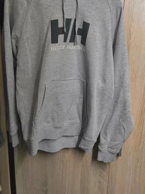 Grå hoodie från Helly Hansen - Säljer en grå hoodie från Helly Hansen med stor HH-logga på bröstet. Hoodien har huva med dragsko och en stor magficka. Materialet är mjukt och bekvämt, perfekt för chill dagar. Klassisk passform och snygg design som funkar till det mesta.
