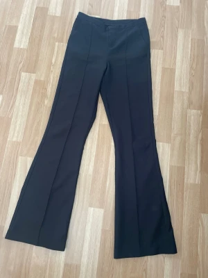 Svarta utsvängda kostymbyxor  - Snygga svarta byxor med utsvängda ben och pressveck framtill. Byxorna har medelhög midja och klassisk passform, perfekta för en trendig look. 