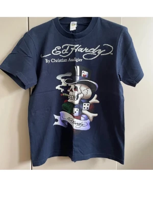 Mörkblå Ed Hardy t-shirt med tryck - Snygg mörkblå t-shirt från Ed Hardy med stort färgglatt tryck av dödskalle, hatt, tärningar och cigarr på framsidan. Klassisk logga både fram och bak. Perfekt för dig som gillar streetwear och unika motiv.