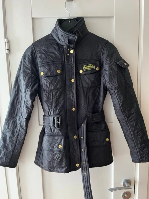 Svart quiltad jacka från Barbour - Snygg svart quiltad jacka från Barbour International med bälte i midjan och gulddetaljer på knapparna. Jackan har hög krage, fyra fickor framtill och ett broderat Barbour-märke på bröstet. Perfekt för dig som vill ha en klassisk och stilren look.
