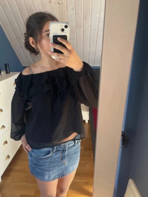 Svart offshoulder blus med volang - Trendig svart offshoulder blus med volangdetaljer längs överdelen. Blusen har långa, transparenta ärmar och är tillverkad i ett lätt och luftigt tyg som ger en snygg och avslappnad look. Perfekt att matcha med jeans eller kjol för en cool stil.