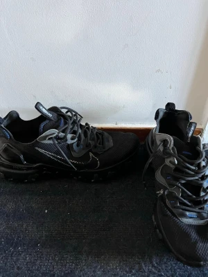 Nike React Element 55 svarta sneakers - Svarta Nike React Element 55 sneakers med mesh-överdel, reflekterande detaljer och synlig vit swoosh på sidan. Skorna har svarta snören, blå accenter och en unik vågig sula för extra grepp. Perfekta för dig som gillar sportig och modern stil. ( inga defekter, box finns inte)