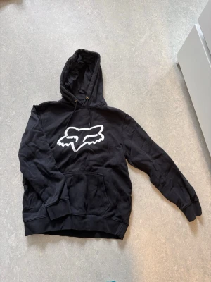 Svart Fox hoodie med logga - Svart hoodie från Fox med stor vit logga framtill. Klassisk modell med huva, dragsko och känguruficka. Mjuk bomullsblandning och ribbade muddar vid ärmslut och nederkant. Perfekt för en avslappnad streetstil.