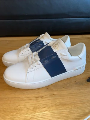 Valentino vita sneakers med blå detalj - Snygga vita sneakers från Valentino med en bred blå läderdetalj över snörningen och ikoniska nitar baktill på hälen. Skorna har en klassisk låg profil och är tillverkade i slätt läder med vita skosnören och diskret logga på plösen.