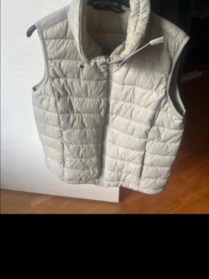 Beige dunväst med hög krage - Snygg beige dunväst med quiltat mönster och hög krage. Västen har dragkedja framtill och två sidofickor. Perfekt för lager-på-lager och ger en clean look. Materialet är dun och syntet, vilket gör den både varm och lätt.