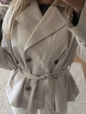 Beige kort trenchcoat med knytskärp - Stilren beige trenchcoat med dubbelknäppning och knytskärp i midjan. Jackan har bred krage och stora knappar framtill. Perfekt för dig som gillar klassisk och tidlös stil. Materialet ser ut att vara bomull eller bomullsblandning. Endast använd en gång! Säljer pga att den är för kort på mig!