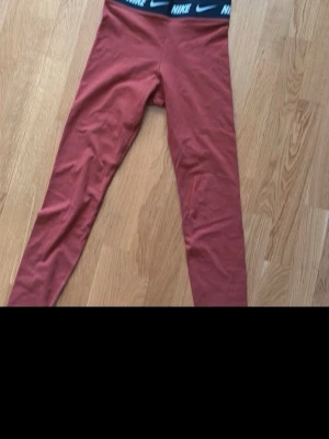 Rostfärgade leggings från Nike - Nike leggings i snygg rostfärg med svart resår i midjan där Nike-loggan syns flera gånger. Tight passform och stretchigt material som sitter skönt. Perfekta för träning eller chill. Klassisk Nike-kvalitet och stil.