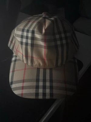 Burberry keps med klassiskt rutmönster - Snygg keps från Burberry med det ikoniska beigea rutmönstret i svart, vitt och rött. Justerbar baktill med svart spänne och Burberry London England-logga. Perfekt accessoar för att lyfta din streetwear-look.