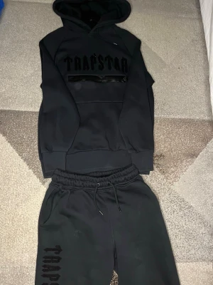 Trapstar tracksuit - Svart hoodie från Trapstar med stor broderad logga på bröstet och klassisk huva med dragsko. Hoodien har en ficka framtill med dragkedja och är gjord i mjukt bomullsmaterial. Perfekt för en streetwear-look.