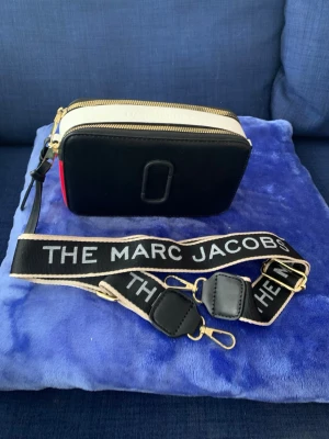 Marc Jacobs Snapshot svart/vit väska - Säljer en the Marc Jacobs Snapshot handväska i svart och vitt med guldfärgade dragkedjor och bred axelrem. Väskan är rektangulär och har detaljer i konstläder samt tydlig logga på ovansidan. Perfekt för dig som gillar trendiga accessoarer. Inköpt utomlands inget kvitto el äkthetsbevis, inga garantier gällande märket säljes därför billigt. Helt oanvänd.