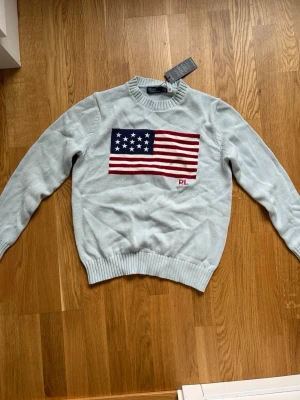 Ljusblå stickad tröja Polo Ralph Lauren - Ljusblå stickad tröja från Polo Ralph Lauren med amerikansk flagga i rött, vitt och blått på bröstet samt RL-broderi. Tröjan har rund halsringning och ribbade muddar vid ärmslut och nederkant. Perfekt för dig som gillar klassisk preppy stil.