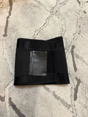 Svart midjetränare shapewear - Svart midjetränare i elastiskt material med bred kardborreknäppning och förstärkta paneler. Perfekt för att forma midjan och ge extra stöd. Materialet är andningsbart och stretchigt för bekväm passform.