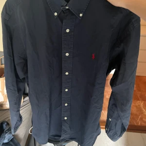 Ralph lauren skjorta  -  Strlk S-M Klassisk mörkblå skjorta från Polo Ralph Lauren i 100% bomull. Skjortan har långa ärmar, knappar framtill och ikonisk röd broderad logga på bröstet. Perfekt för dig som gillar stilrena och tidlösa plagg med snygga detaljer. Den är stor i storleken. Passar för M eller L