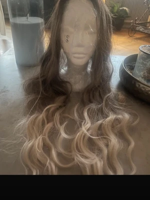Lång vågig ombre peruk - Snygg lång peruk med mjuka vågor och ombre-effekt från mörkbrun botten till ljusblonda toppar. Perfekt för dig som vill ha en naturlig look med extra volym och längd. Materialet ser ut att vara syntetiskt hår.