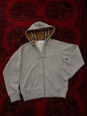 Grå hoodie från Burberry med dragkedja - Snygg grå hoodie från Burberry med klassiskt rutigt foder i huvan. Tröjan har dragkedja framtill, två fickor och ribbade muddar. Tillverkad i 100% bomull för en skön och mjuk känsla. Perfekt för en chill och stilren look.
