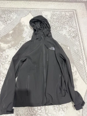 Svart vindjacka från The North Face - Säljer en svart vindjacka från The North Face med huva och dragkedja framtill. Jackan har två fickor med dragkedja och justerbar huva, perfekt för blåsiga dagar. Klassisk logga på bröstet och ryggen. Har använt den 1 gång. Jag fick den som present av en vän och den var liten på mig. Kan diskutera om priset