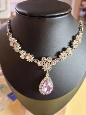 Silverfärgat halsband med lila sten - Ett glittrande halsband i silverfärg med blommiga detaljer av klara stenar och en stor droppformad lila sten som hänge. Halsbandet har en elegant och lyxig känsla med många gnistrande stenar som fångar ljuset.