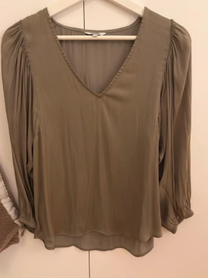 Beige blus med v-ringning från mbyM - Stilren beige blus från mbyM med v-ringad hals och långa, lätt puffade ärmar. Tillverkad i ett tunt, flowy material som ger en snygg siluett. 