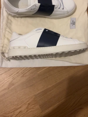 Valentino vita sneakers med blå detalj - Snygga vita sneakers från Valentino med en bred mörkblå läderdetalj över snörningen. Skorna har klassisk låg profil, vita skosnören och diskret logga på plösen. Tillverkade i slätt läder för en clean och exklusiv look. Endast skorna tillkommer 
