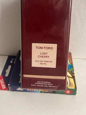 Tom Ford Lost Cherry EdP 100 ml - Tom Ford Lost Cherry Eau de Parfum 100 ml är en lyxig parfym i en elegant mörkröd glasflaska. Doften är känd för sina söta och fruktiga toner med inslag av körsbär och kryddor. Flaskan har en stilren design med en beige etikett framtill.