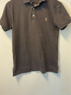Mörkgrå pikétröja från Ralph Lauren - Klassisk mörkgrå pikétröja från Ralph Lauren med korta ärmar och krage. Tröjan har knappar framtill och den ikoniska broderade loggan på bröstet. Perfekt för en stilren och avslappnad look.