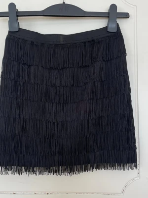 Svart franskjol med elastisk midja - svart kjol med flera lager fransar som ger en snygg och rörlig look. Kjolen har elastisk midja och är kort i modellen. Materialet känns mjukt och följsamt mot kroppen.