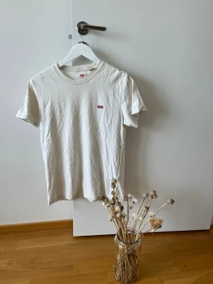 Vit t-shirt från Levi's med logga - Klassisk vit t-shirt från Levi's med den ikoniska röda loggan på bröstet. T-shirten har rund halsringning och korta ärmar. Perfekt basplagg i mjuk bomull som funkar till allt.