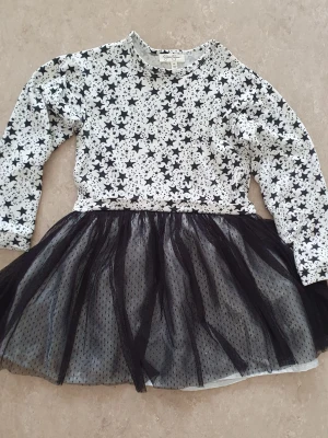 Klänning - Superfin kort klänning med svartvit stjärnmönstrad överdel och fluffig svart tyllkjol. Långärmad och rund halsringning, perfekt för dig som gillar en lekfull och cool stil. Materialet känns mjukt och bekvämt mot huden.
