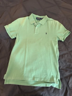 Grön pikétröja från Polo Ralph Lauren - Snygg ljusgrön pikétröja från Polo Ralph Lauren med klassisk krage, korta ärmar och broderad logga på bröstet. Slim fit-modell i mjuk bomull, perfekt för en avslappnad och stilren look. Storlek 14-16