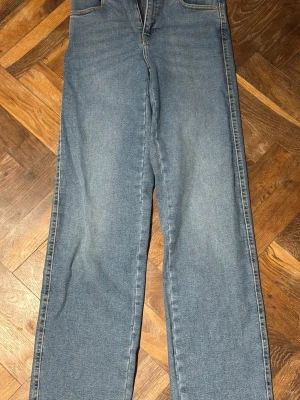 Blå raka jeans i klassisk modell - Säljer ett par blå jeans med rak passform och hög midja. Jeansen har klassisk femficksdesign och är tillverkade i denim. Perfekta för en avslappnad och stilren look.